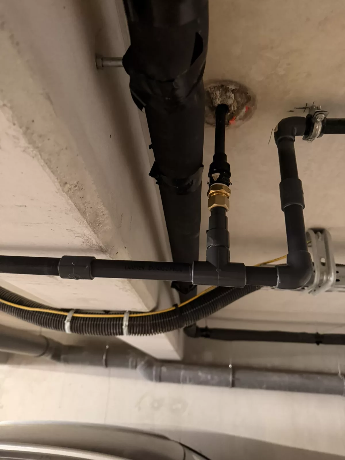 Vue du plafond technique du parking montrant le raccordement en PVC Pression 25 réparé, avec té et jonction laiton reliant le nouveau tronçon à l'attente en multicouche existante.