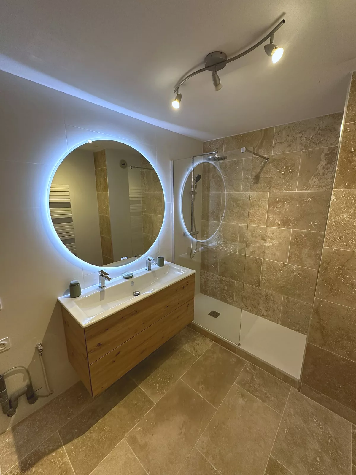 Douche à l'italienne en travertin véritable avec meuble double vasque et miroir rond rétroéclairé, salle de bain rénovée à Toulon.