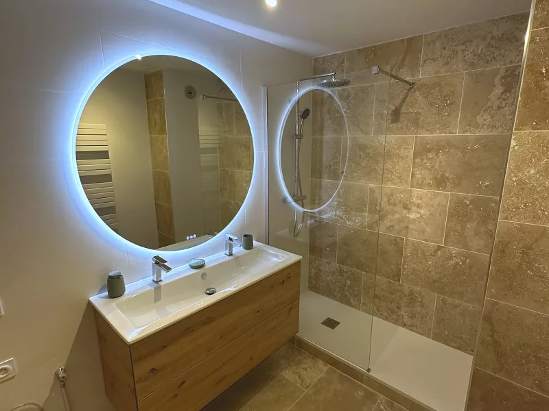 Douche à l'italienne en travertin véritable avec meuble double vasque et miroir rond rétroéclairé, salle de bain rénovée à Toulon.