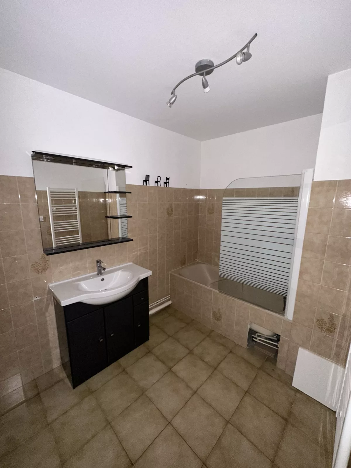 Salle de bain vétuste avant rénovation : ancien carrelage beige, baignoire avec pare-douche et meuble vasque à remplacer.