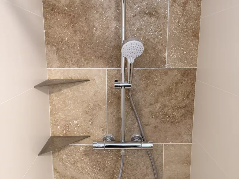 Détail du mitigeur thermostatique et du flexible de douche sur fond de travertin.