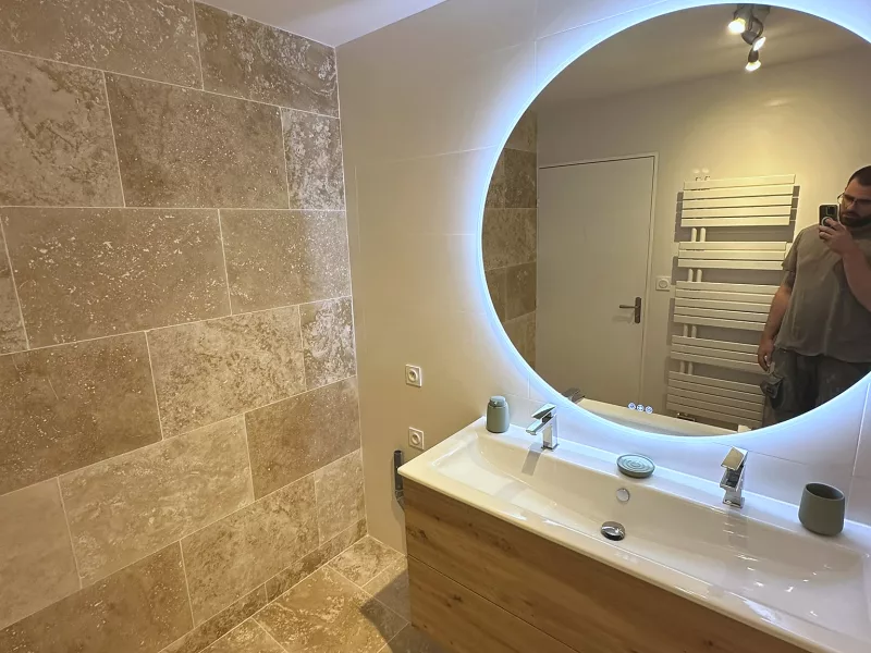 Vue d'ensemble de la salle de bain rénovée à Toulon : double vasque, douche à l'italienne en travertin et carrelage blanc.